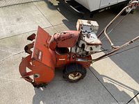 Front-left view of the orange metal housing and chute with white engine housing showing surfaces rust and wear, tire with chains visible