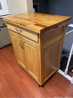 Kitchen island cart viewed diagonally showing hardwood top, MDF doors, drawer, side towel holder, and caster wheels.