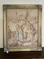 Frontal photo of large framed tapestry showing historical English scene with figures and a dog, boats in background
