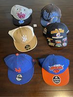 Photo showing six sports hats on a table including Knicks, Orlando Magic, New York Mets, Phoenix Suns, Pittsburgh Steelers, and a tan and white New York fitted hat.