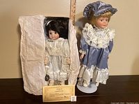 Photo showing both porcelain dolls side by side with a ruler behind for size reference, certificate of authenticity visible near the smaller doll wrapped in tissue.