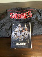 Back of black vintage Reebok jacket showing SABRES in large red/white letters with red piping, laid flat on table with yearbook in front