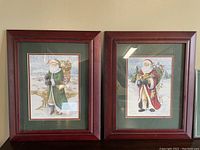 Two framed watercolor prints of Santa Claus by Susan Baldwin placed side by side, showing the overall presentation including the cherry wood frames and green mats.