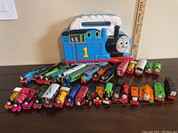 Photo showing the Thomas-shaped carrying case with 33 assorted die-cast trains and construction equipment pieces arranged in front, displaying a colorful collection of engines, cars, and trucks.
