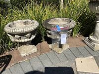 Two matching heavy garden planters shown side-by-side on a brick and stone patio near tall grass and garden plants.