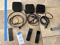 Three Apple TV HD units lined up with three power cables and three remote controls on a tiled floor.