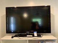 Front view of the Samsung TV powered off on a white table showing the screen, stand and remote placed next to it.