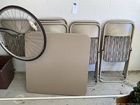 Photo showing two beige rectangular folding card tables leaning against the wall with eight beige folding chairs beside them, some stacked and some standing upright.