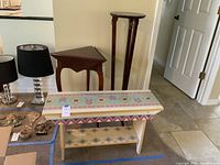 Full view of all three decorative furniture pieces placed in a room: the hand painted bench foreground, wooden night stand table and tall plant stand with lion heads in the background.