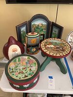 Entire set including hand painted stool, Santa gourd, round box with lid, metal pitcher, and tri-fold decorative screen on white table.