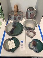 Full set of stoneware pottery showing pitcher, basket with woven handle, pie plate, scalloped bowl, heart-shaped dishes, and small dish with spout. Variations of green, brown and purple glaze visible.