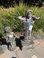 Two stone statue garden decorations, one smaller girl statue with hands on hips, and one larger girl statue holding two bowls and standing on a decorative base.