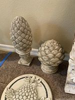 Two white pineapple statues and round welcome plaque arranged on carpet against wall.