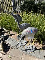 Three metal sandhill crane statues in backyard landscaping, showing one feeding pose on round base, two standing poses with round and grass-like bases