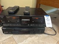 Sony VHS player and stereo cassette tape deck side by side with remotes on top, showing front control panels and buttons.