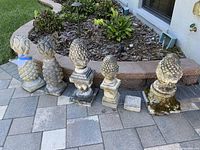 Wide shot showing five stone pineapple statues lined up on paved patio near garden bed.