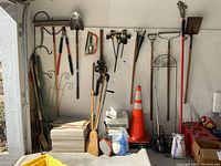 Full view showing an assortment of long-handled yard tools, clamps, traffic cones, bags of garden supplies, and a stack of tiles or pavers.