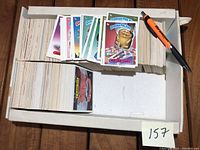 White storage box containing loose stacks of Garbage Pail Kids cards, with pen for scale and note '157'.