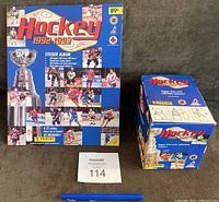 Photo of the sticker album and the unopened box of sticker packs, placed side by side with a pen for size reference.