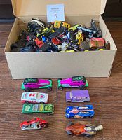 Photo showing a box full of assorted diecast vehicles piled inside with several vehicles laid out outside the box on a wood floor.