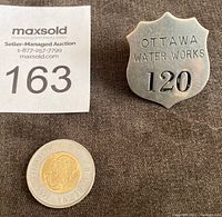 Front view of the shield-shaped metal badge alongside a Canadian two-dollar coin for scale.