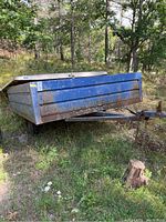 Blue metal utility trailer from a frontal-side angle showing rust and paint wear, with lid partially off.