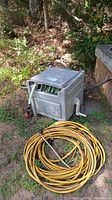 Garden hose reel box with green hose and yellow heavy duty extension cord coiled beside it on the ground outdoors.