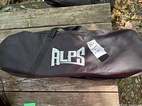 Closed black ALPS carrying case with handles lying on wooden surface outdoors, showing ALPS logo on side.