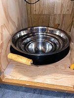 Nested stainless steel bowls inside a black wok with wooden handles, displayed inside wooden shelving.