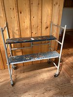 Front view of small scaffold showing two wooden plank steps with paint splatters and metal frame with four caster wheels.