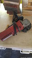 Red Milwaukee angle grinder placed on a table under bricks, showing side and power cord.