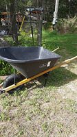 Full side view of a black plastic tub landscape wheelbarrow with wood handles, single black wheel, on grass with trees in background.