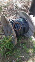 Wide side-view of large wooden spool fully loaded with black tubing, positioned outdoors on grass and leaves.