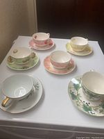 Photo showing six assorted tea cups and saucers in different floral patterns and pastel colors, placed on white surface.