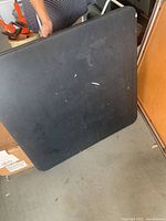 Black folding card table resting on its side, shows scratches and wear on surface.