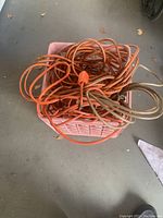Plastic crate filled with loosely coiled orange and beige extension cords on a concrete floor.