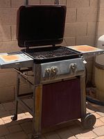 Gas BBQ grill with maroon lid open, showing grill grate and metal frame with side shelves. Visible rust and dirt on metal surfaces.