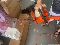 Side view of an orange and black hedge trimmer showing dual-sided cutting blades and ergonomic handles, held by a person's hand.