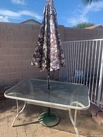 Photo showing the rectangular glass top table with white metal frame and attached patterned fabric umbrella in folded position.