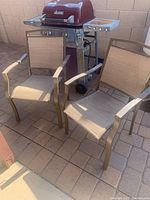Two beige outdoor mesh sling chairs with bronze-colored metal frames placed near a red grill and brick wall.