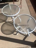 Two round glass top patio side tables seen from above with shadow cast on concrete ground.