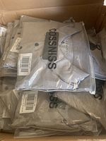 View of multiple men's shirts stacked inside a box. Shirts are grey/khaki, sealed individually in plastic packaging showing the 'Cossniss' brand printed on the package.