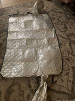 Silver reflective snow/solar cover unfolded on a patterned carpet, showing black edging and securing straps.