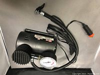 Black portable auto air compressor with built-in analog pressure gauge, attached 12V power cord with cigarette lighter plug, and cooling vents visible on the side.