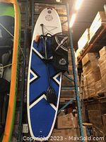Inflated white and blue SUP board leaning against warehouse shelving with paddle, backpack, leash and pump attached.
