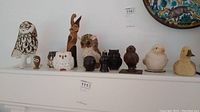 Full view of all bird figurines lined up on a white shelf including wooden, ceramic, and iron pieces