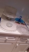Clear glass cake plate with ribbed cover, blue handled serving plate, clear glass devilled egg platter, and clear serving plates on white counter surface.