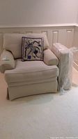 Beige chair with embroidered pillow on seat and extra seat cushion wrapped in plastic beside chair