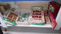 Multiple boxes of Christmas tree ornaments in gold, green, silver, and red colors shown on a white shelf.