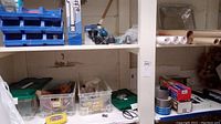 Shelf with blue plastic organizational bins containing small metal screws and fasteners, paper towels and duct tape on the lower shelf, and rolled shelf paper to the right